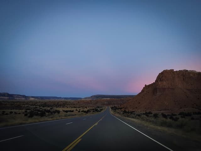 Two-lane highway stretching through a desert landscape toward a mesa at dusk