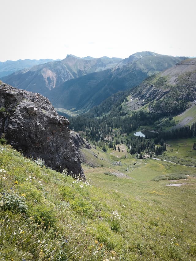 High-elevation mountain valley with rocky slopes, green meadows, scattered trees, and distant ridgeline under a pale sky