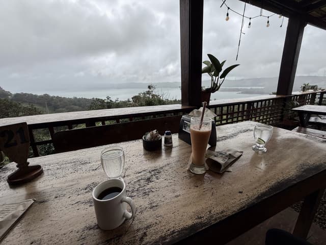 Rainy coastal view from a rustic open-air café, with coffee cups and a smoothie on a weathered wooden table overlooking the misty ocean and lush greenery