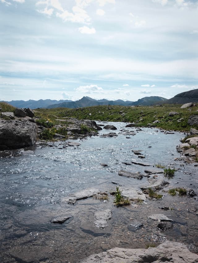 Shallow rocky stream flowing through an open grassy landscape with distant mountains under a cloudy sky