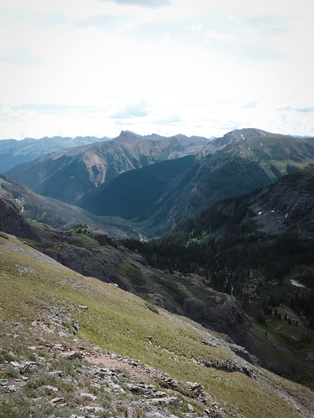 Sunlit alpine valley with rugged, layered mountain ridges and grassy foreground slopes under a hazy sky