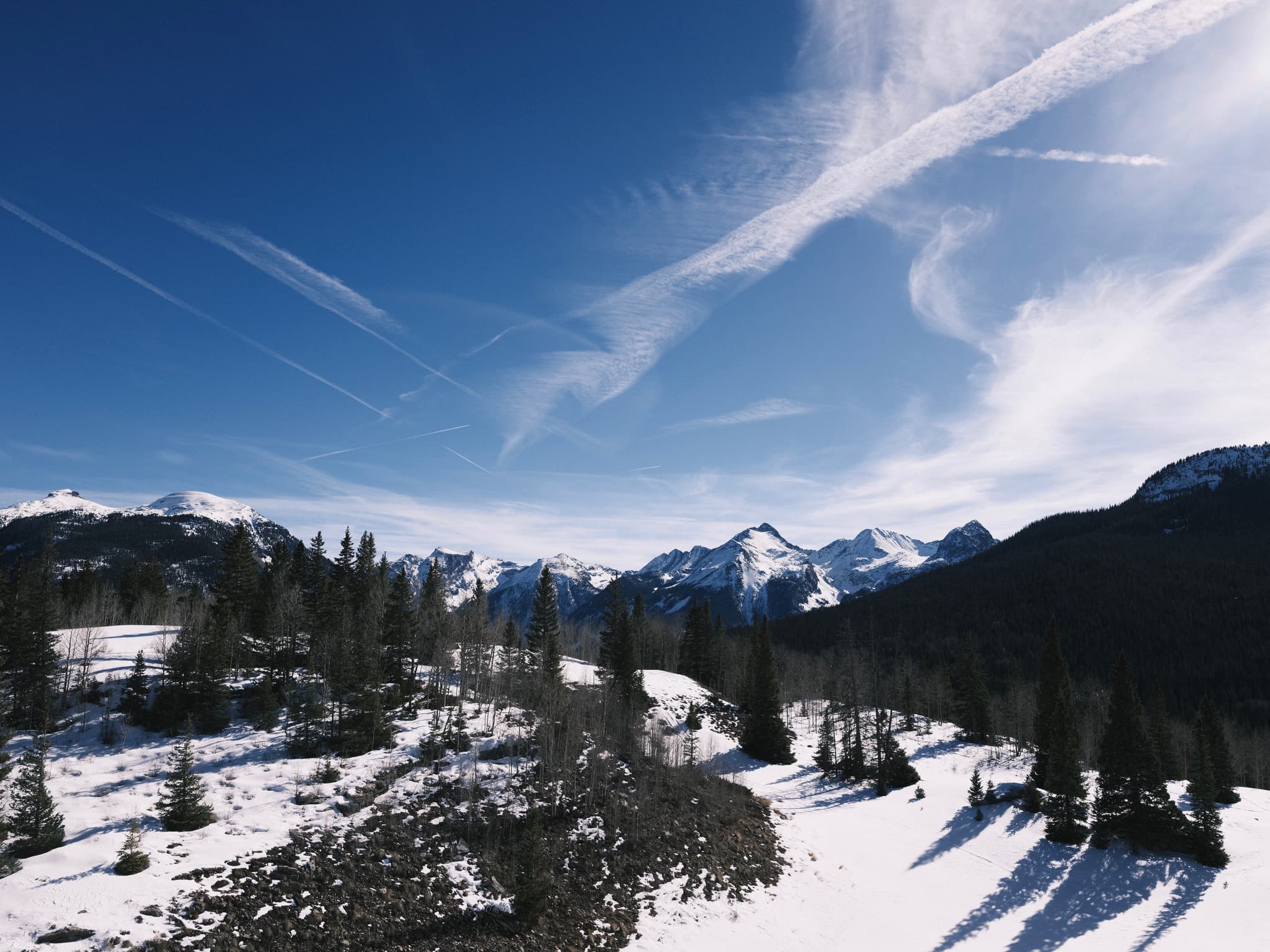 Snow-covered mountain valley with scattered evergreen trees beneath a blue sky with wispy clouds
